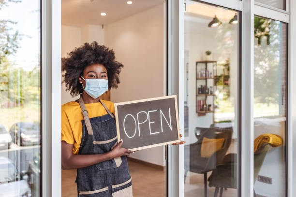 Come one, come all One African-American Woman with surgical protective mask  holding open sign in front of her small business store. open stock pictures, royalty-free photos & images