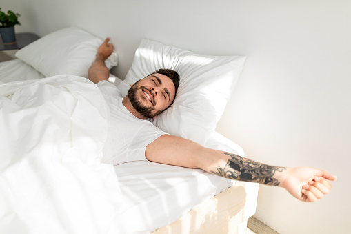 Comfortable sleep. Wellslept tattooed man waking up in morning and stretching hands, smiling with closed eyes while lying in bed, free space. Good morning concept
