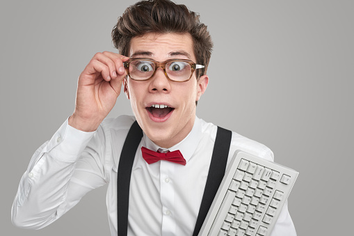 Funny clever male programmer in glasses and with white keypad looking at camera with astonishment on gray background in studio