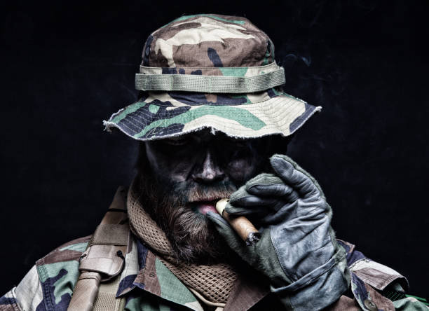 Commando soldier in boonie hat smoking cigar Brutal and serious commando soldier, army special forces veteran, in camouflage battle uniform, boonie hat, black paint on bearded face, combat knife in shoulder holder, smoking cigar, studio portrait special forces stock pictures, royalty-free photos & images