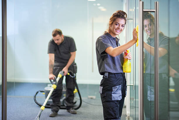 commercial cleaning contractors a female cleaning contractor is polishing the glass partition offices whilst In the background a male colleague steam cleans an office carpet in a empty office in between tenants.  .The female is smiling to camera. cleaner stock pictures, royalty-free photos & images