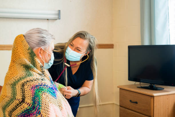 Compassionate nursing care A blond nurse wearing dark navy scrubs and face mask uses her stethoscope to  listen to the lungs of a patient in a medical facility, Midwest, USA inpatient stock pictures, royalty-free photos & images