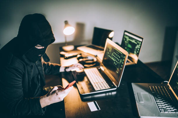 Computer hacker using phone Computer hacker sitting in front of laptop late at night and using phone ski mask criminal stock pictures, royalty-free photos & images