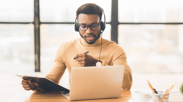 Concentrated african american guy in headset writing notes Conference Call. Focused black man in headset making notes talking at video chat, support agent consulting customer cross section stock pictures, royalty-free photos & images
