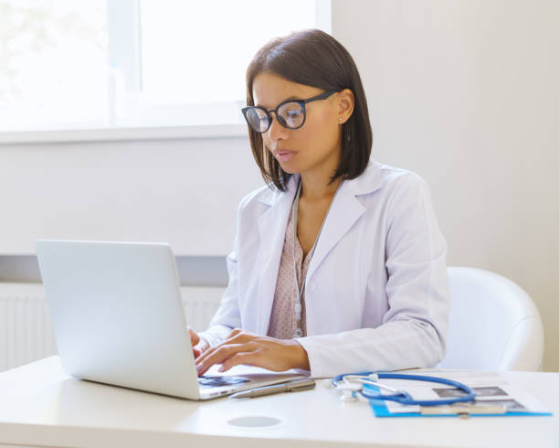 Concentrated afro american woman doctor using laptop while sitting at workplace in medical clinic stock photo