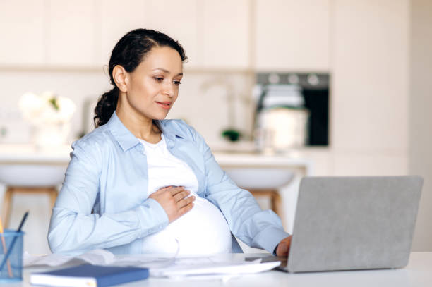 Concentrated beautiful successful mixed race pregnant woman, freelancer, manager or designer working remotely at laptop, checking business report or project design, stroking belly, smiling stock photo