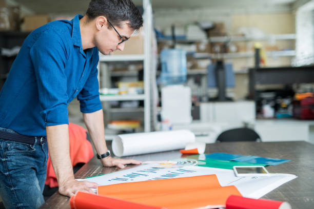 Concentrated designer thinking about paper for printing Serious concentrated male designer in blue shirt standing at table and analyzing quality of banner while thinking about paper for printing in printing house printing plant stock pictures, royalty-free photos & images