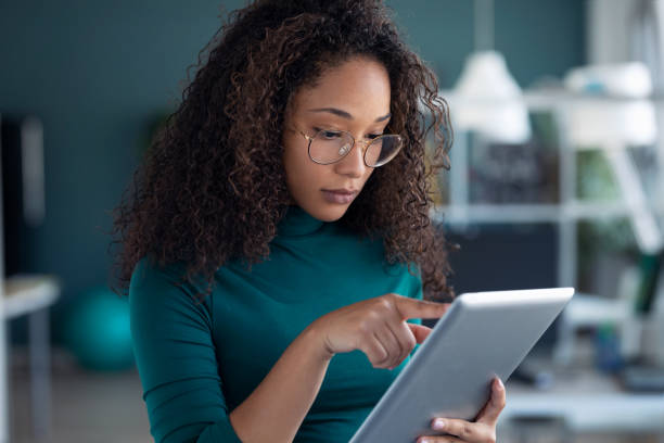 Concentrated young entrepreneur woman using her digital tablet while standing in the office. Portrait of concentrated young entrepreneur woman using her digital tablet while standing in the office. using digital tablet stock pictures, royalty-free photos & images