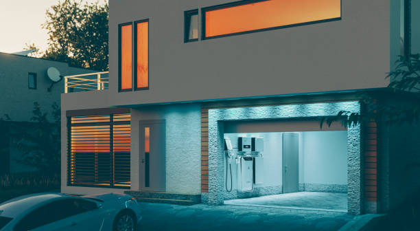 concept of a home battery energy storage system located in the garage of a modern family house in a futuristic blue light illuminating the evening atmosphere of a quiet street. 3d rendering. stock photo
