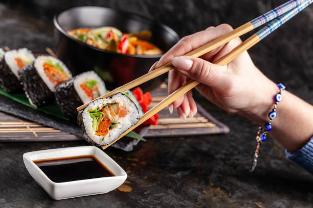Concept of Asian cuisine. The girl is in a Chinese or Japanese restaurant sushi, holds wooden sticks in his hands. Dunk sushi in soy sauce. Different Asian dishes are on the table stock photo