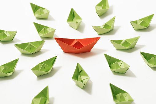 Orange paper boat among of a group of green paper boats on white background. Concept of leadership, individuality, unique.