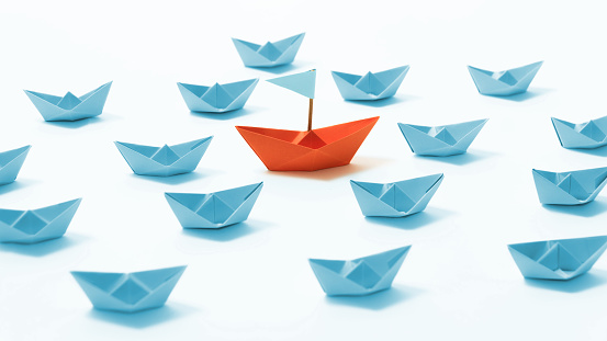 Orange paper boat among of a group of blue paper boats on white background. Concept of leadership, individuality, unique.
