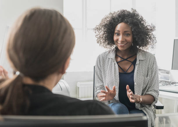 Confident African businesswoman with Afro hairstyle gesturing and smiling towards male colleague Attractive businesswoman in her 30sin office with co-worker, talking with cheerful expression assertiveness stock pictures, royalty-free photos & images