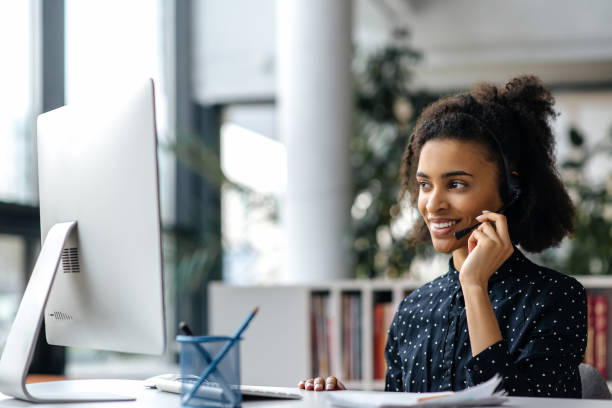 Confident beautiful african american girl in headset, manager, call center worker or freelancer, sitting at the table, using computer, communicating with colleague or client via video link, smiling stock photo