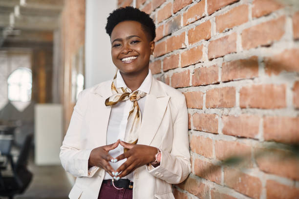 Confident black businesswoman leaning on a wall in the office and looking at camera. stock photo