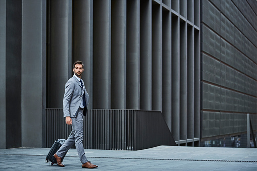 Full length of confident businessman with luggage. Side view of executive is walking against building. Professional is looking away.