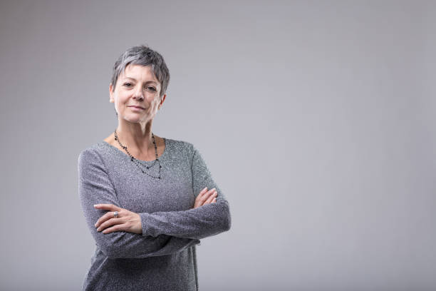 Confident businesswoman with folded arms Confident businesswoman with folded arms standing looking intently at the camera over grey with copy space senior women stock pictures, royalty-free photos & images