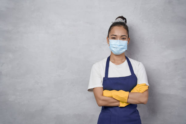 Confident cleaning lady wearing rubber gloves and medical protective face mask keeping arms crossed, looking at camera while standing against grey wall Confident cleaning lady wearing rubber gloves and medical protective face mask keeping arms crossed, looking at camera while standing against grey wall. Cleaning services during covid 19 pandemic maid stock pictures, royalty-free photos & images