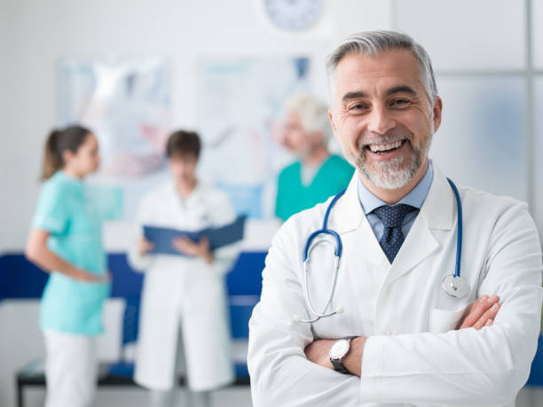 Confident doctor posing at the hospital Confident smiling doctor posing and the hospital with arms crossed and medical team working on the background general practitioner photos stock pictures, royalty-free photos & images