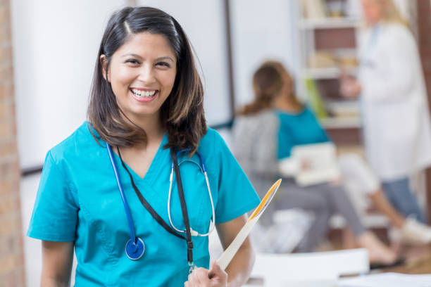 Confident Filipino nurse at work in emergency room Smiling mid adult Filipino nurse holds a patient's medical chart in emergency room. A doctor is with a patient in the background. filipino ethnicity stock pictures, royalty-free photos & images