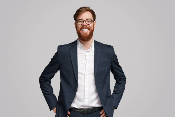 Confident ginger businessman smiling for camera Successful Irish businessman with ginger beard cheerfully smiling and looking at camera while standing against gray background business suit stock pictures, royalty-free photos & images