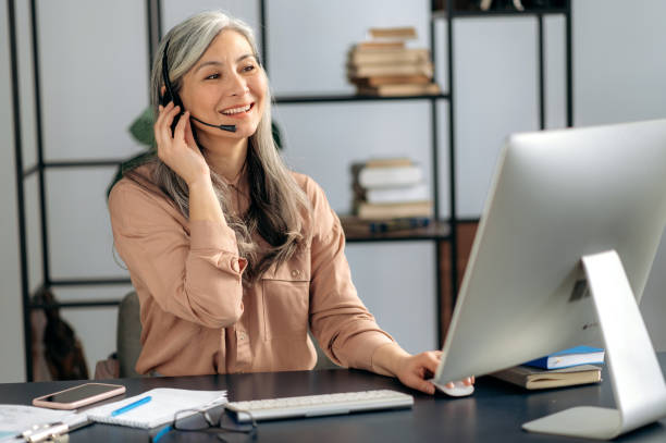 Confident influential senior Asian woman, business lady, manager, call center operator, works in office, communicates with client via video call, conducts online consultation, uses headset, smiles stock photo
