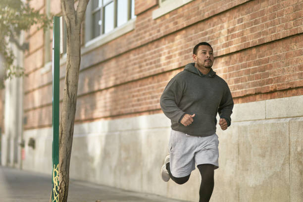 Confident male athlete jogging on sidewalk in city Confident male athlete jogging on sidewalk by wall. Full length of determined young man is exercising in city. He is in sportswear. body positive stock pictures, royalty-free photos & images