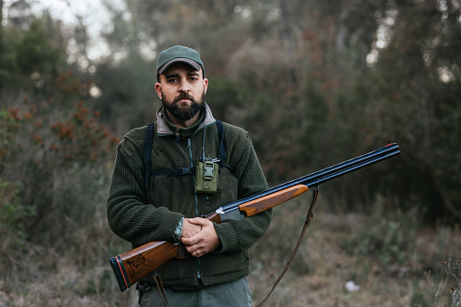 Serious bearded male hunter in outerwear standing with rifle for hunting among plants in woods and looking at camera