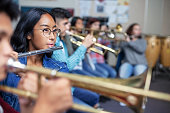 Confident mixed ethnicity Asian teenage girl playing flute in musical orchestra band class. Students practicing music in high school. She is learning woodwind instrument.