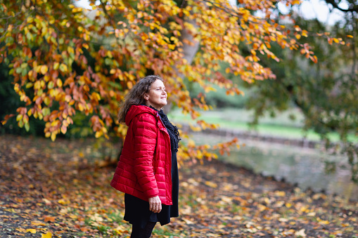 Senior woman with beautiful long curly hair,wearing red jacket and black dress and scarf, standing in autumnal park
