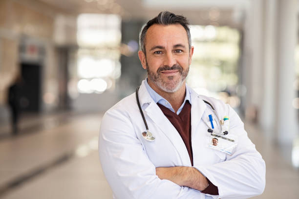 Confident successful mature doctor at hospital Portrait of happy doctor in labcoat and stethoscope standing with folded hands in modern hospital corridor. Successful general practitioner looking at camera with satisfaction at private clinic. Confident proud head physician standing in hospital hallway smiling with copy space. medical field stock pictures, royalty-free photos & images