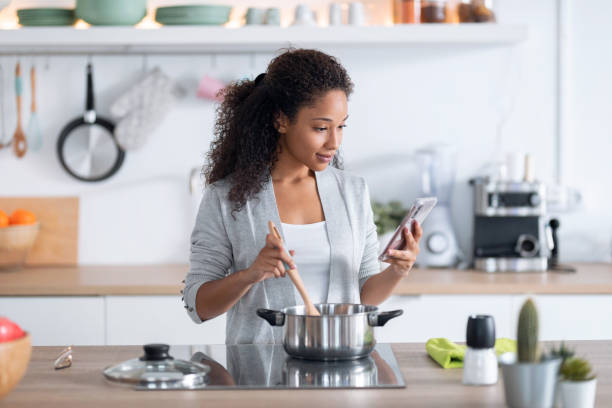 zekere jonge afrikaanse amerikaanse vrouw gezond voedsel terwijl het gebruiken van haar mobiele telefoon in de keuken thuis. - afro amerikaanse etniciteit stockfoto's en -beelden
