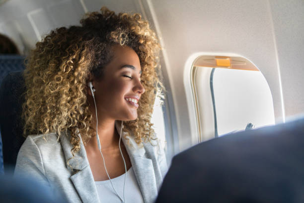 Confident young woman enjoys flight Attractive young woman smiles while listening to musing during a long flight. She is sitting next to the window. plane window seat stock pictures, royalty-free photos & images