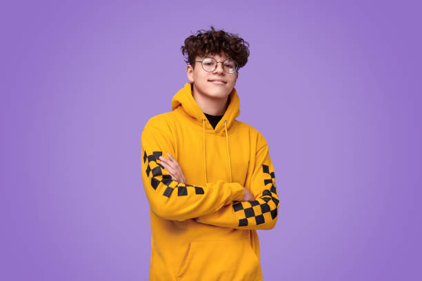 Confident youngster looking at camera Positive teen boy in yellow hoodie crossing arms and looking at camera against bright violet background teenagers stock pictures, royalty-free photos & images