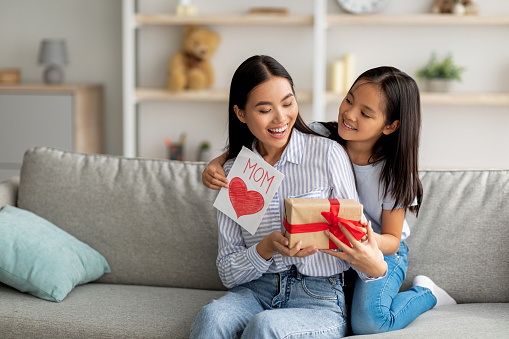 Congratulations. Loving asian daughter greeting surprised mom with Mother's Day, giving her handmade card and gift box, sitting on sofa at home interior, free space
