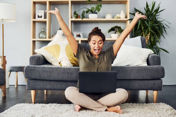 Congratulations! You're the winner! Shot of a young woman cheering while using a laptop on the living room floor at home good news stock pictures, royalty-free photos & images