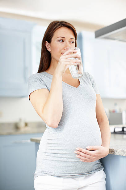 Conscientious about the health of her baby A smiling pregnant woman drinking water in the kitchen looking thoughtful hydration during pregnancy stock pictures, royalty-free photos & images