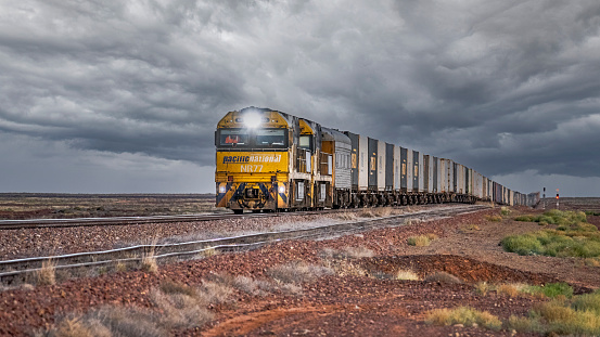 Pimba, Australia - March 13, 2022: Pacific National intermodal container freight train heading east across sparse desert under stormy skies.  Metaphor for business in trouble. Image manipulation - incorporates  sky replacement.