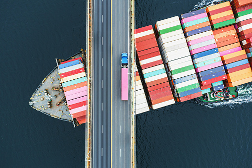 Aerial view of a container ship passing beneath a suspension bridge. Semi truck  with pink cargo container crosses above.