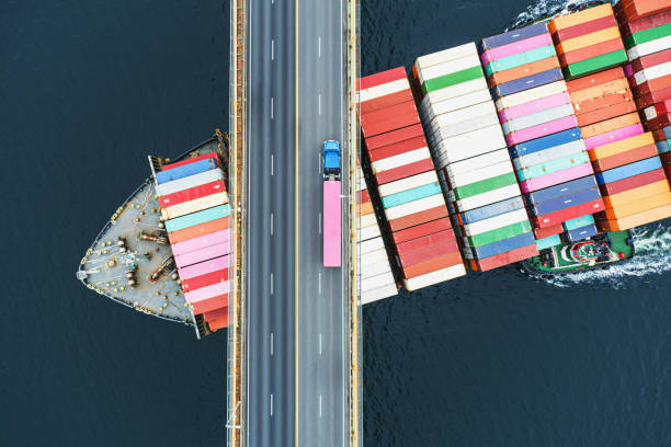 Container Ship Beneath Bridge Aerial view of a container ship passing beneath a suspension bridge. Semi truck  with pink cargo container crosses above. container photos stock pictures, royalty-free photos & images