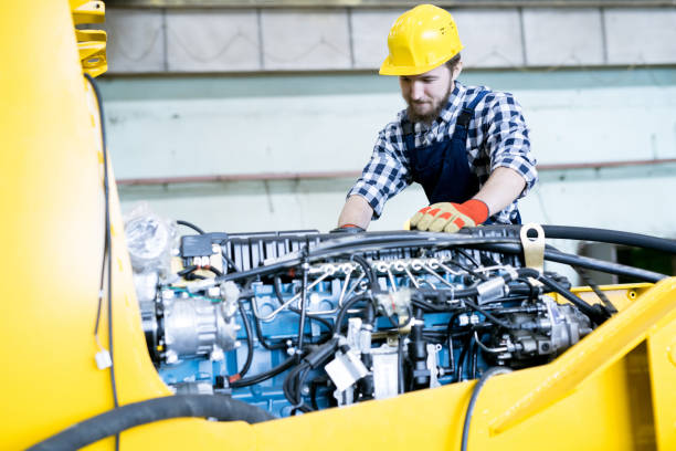 Content concentrated young bearded repairing worker in yellow hardhat examining engine of tractor while working at industrial agricultural machinery plant Repairing worker examining engine of tractor agricultural machinery stock pictures, royalty-free photos & images