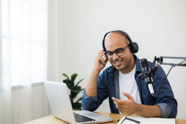 Content creator asia man host streaming his a podcast on laptop with headphones and condenser microphone interview guest conversation at home broadcast studio. Male blogger recording voice over radio stock photo