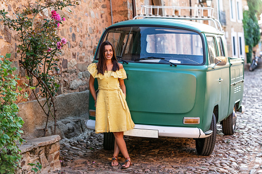 Content female in summer wear standing near vintage van on cozy narrow street of old town and looking at camera. Vacation and travel concept