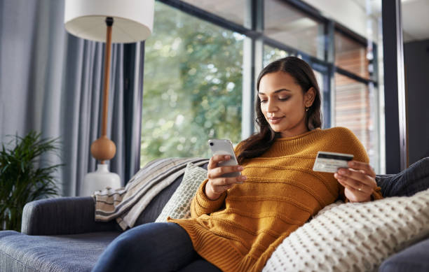 Convenience at it's best Shot of a beautiful young woman using her cellphone and credit card while relaxing on a couch at home online shopping stock pictures, royalty-free photos & images