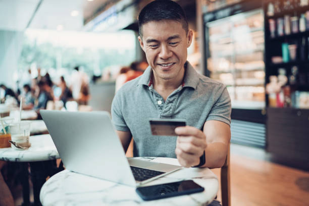Convenient on-line shopping Smiling mid adult Asian ethnicity  man with laptop and credit card in cafe credit card purchase stock pictures, royalty-free photos & images