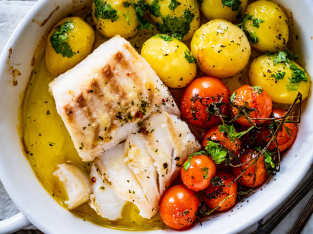 Cooked cod steak, potatoes and cherry tomatoes served in ceramic pan on wooden table stock photo