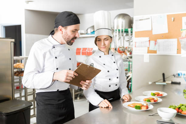 Cooks Going Through Checklist In Kitchen Male and female chefs discussing over clipboard while preparing dish in kitchen menu review stock pictures, royalty-free photos & images