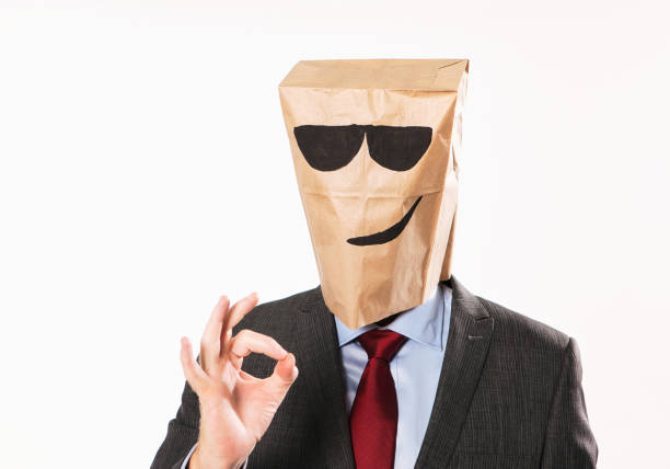 Cool businessman in suit, wearing cartoon paper bag, looks smug as he makes the OK sign with his hand stock photo