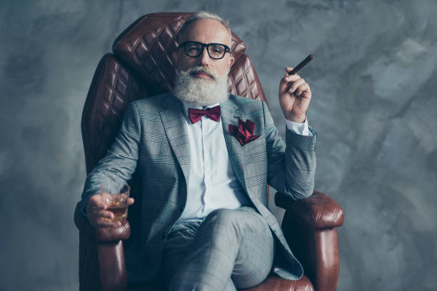 Cool man in glasses, hold cigarette,  glass with brandy, in formal wear, tux with red bowtie and pocket square, sit in leather chair over gray background, looking to the camera, shares, stock, money Cool man in glasses, hold cigarette,  glass with brandy, in formal wear, tux with red bowtie and pocket square, sit in leather chair over gray background, looking to the camera, shares, stock, money snob stock pictures, royalty-free photos & images