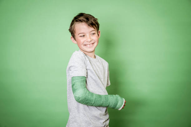 cool young schoolboy with broken arm and green plaster standing in front of green background stock photo